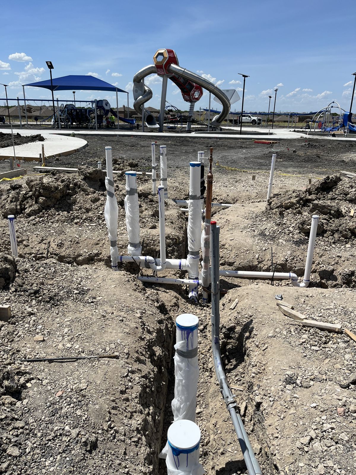 Water-feature plumbing rough-in at a municipal splash pad with playground in the background